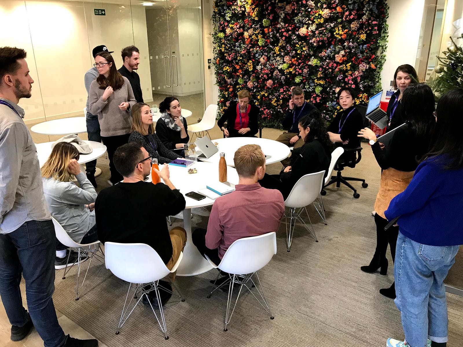 About fifteen designers gathered around tables in an office kitchen with a wall of flowers behind them