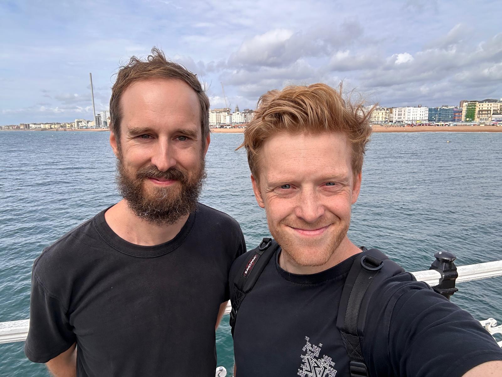 Tom and Jonny smiling in a selfie on Brighton Pier, with the sea and the seafront behind them