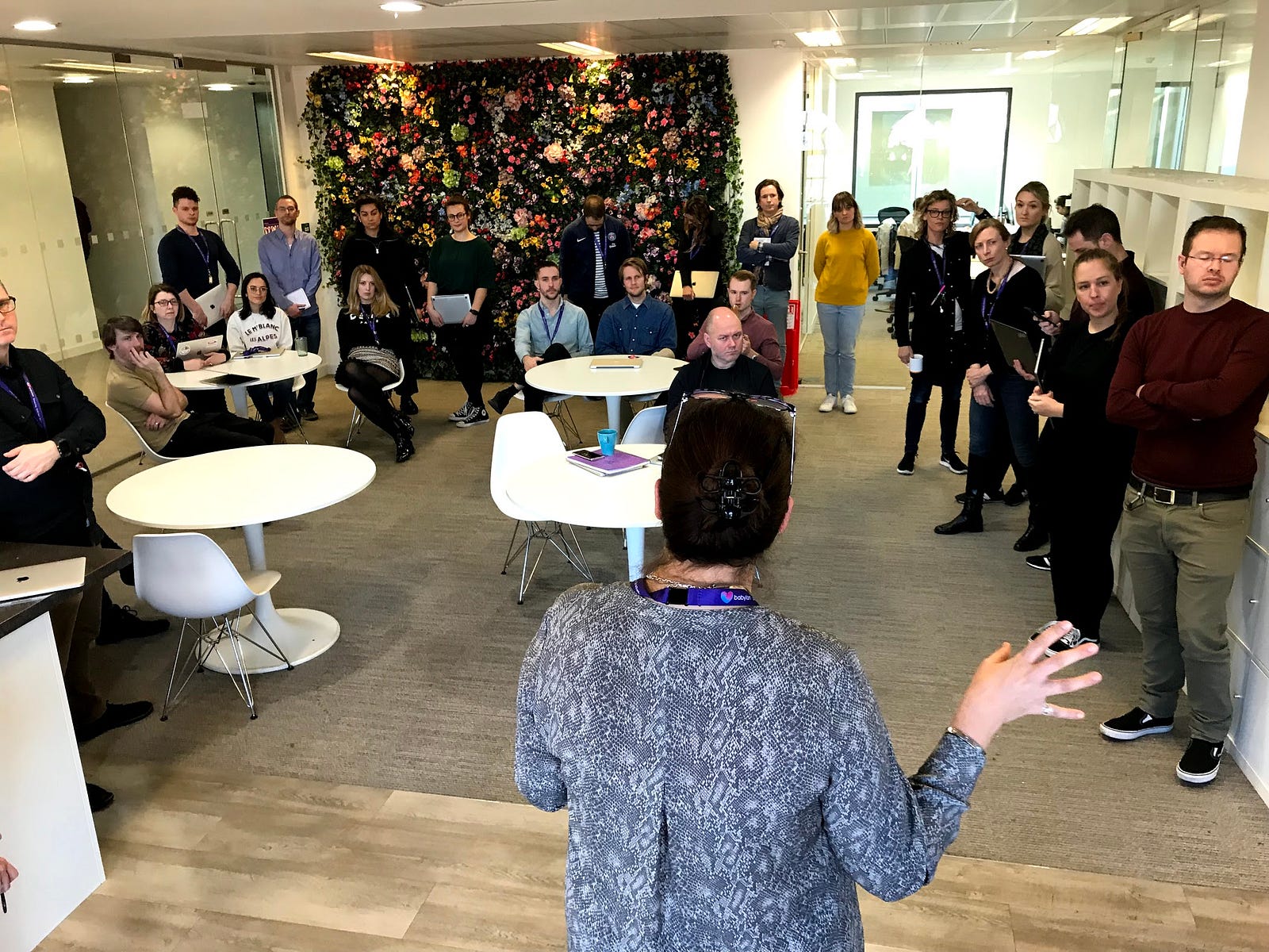 A larger group of around thirty designers filling the same office kitchen in front of the flower wall, listening to a speaker