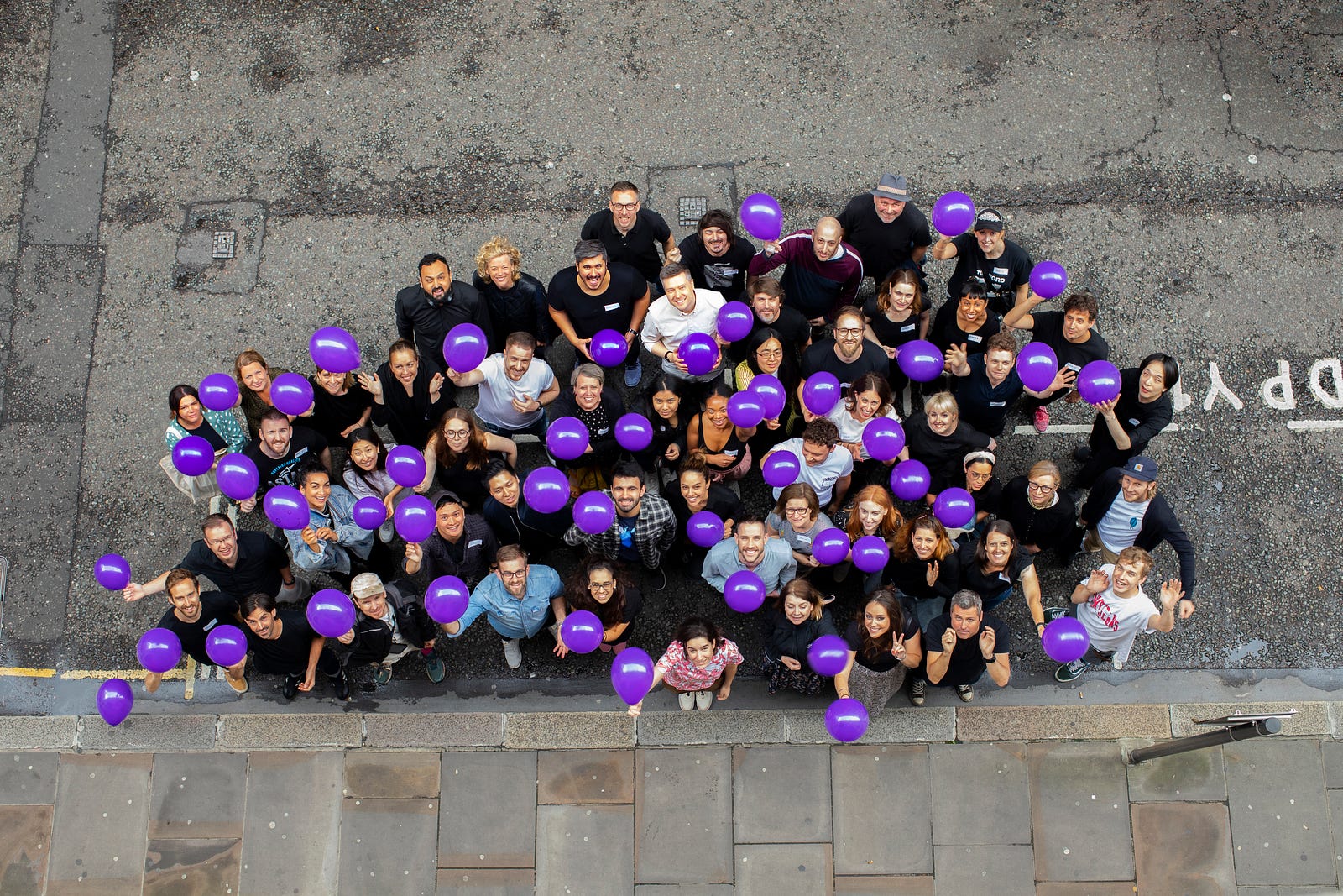 Overhead photo of around 60 members of Babylon’s Experience Design team standing in a street, smiling up at the camera and holding purple balloons